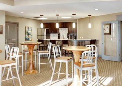 The resident lounge at The Station at Lyndhurst Apartments featuring high-top wooden tables with matching barstools, a modern kitchenette with stainless steel appliances, cabinetry, and pendant lighting.