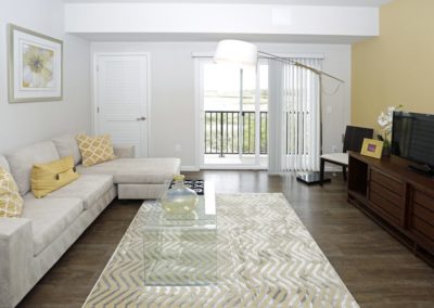 A living room at The Station at Lyndhurst Apartments featuring a sectional sofa, a glass coffee table, and an area rug. Natural light emits from sliding glass doors leading to a balcony.