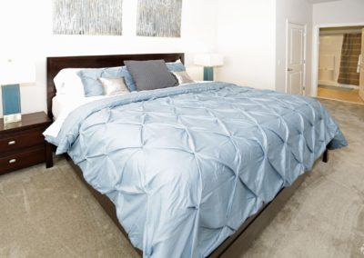 A bedroom at The Station at Lyndhurst Apartments featuring a king-sized bed and carpeting with an en-suite bathroom visible in the background.